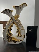 Front angle showing the white ceramic vase with gold accents and detailed floral and grape design.