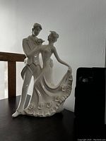 Photo of a white ceramic bride and groom figurine on a table with wood chair visible behind and a dark object to the right.