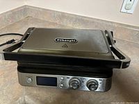 Photo showing the large Delonghi grill with stainless steel top, digital display and two control knobs, placed on kitchen counter.
