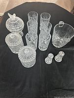 Set of clear cut glassware and serving pieces displayed on a black table surface showing detailed cut patterns and various shapes.