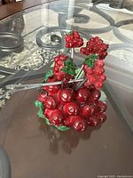 Four small metal fruit forks featuring handles designed as red grape clusters with green leaves, displayed on a glass surface.