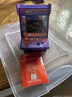 Photo showing purple My Arcade Pocket Player handheld retro game with screen, controls, instruction manual, and clear plastic case on wooden surface.