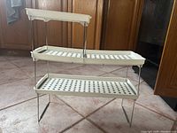Multi-tiered kitchen storage rack shown from the front and angled side, displaying perforated off-white plastic trays and metal legs.