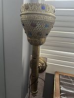 Front view of a brass decorative lamp with intricate filigree work and embedded colorful glass stones. Visible electric cord and a wall mounting apparatus.