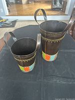 Photo showing two cylindrical metal vases with one handle each, featuring colorful geometric patterns at the base and ornate decoration around the rim, placed on a black cloth.
