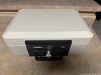 Photo of grey SentrySafe safe box model 1160 with black front panel, key lock, and handle on a stone surface.