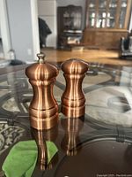 Set of two copper salt and pepper shakers placed on a glass table in an indoor setting.