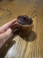 Small brown ceramic pot with lid placed on a wooden surface with a hand for scale.