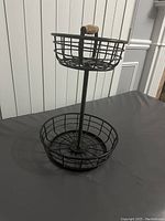 Two-tiered black metal wire basket with a wooden handle on top, shown on a black surface against a white wall.