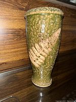Photo of a small green ceramic vase with beige embossed leaf pattern, standing on a reflective dark surface with wooden background.