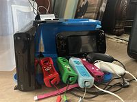 Black Wii U console, four silicone-sleeved Wii Remotes, GamePad controller, and two Nunchuks with cables on floor.