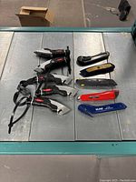 Photo showing ten box cutters on a metal surface, including five black Uline folding knives with lanyards and five assorted rigid cutters.