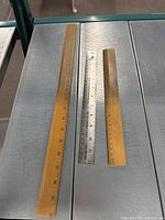 Photo of three rulers displayed on metal surface, showing a long wooden ruler, a medium metal ruler, and a shorter wooden ruler.