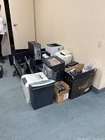 Photo showing printers, monitors, and desktop tower gathered against a beige wall on carpeted floor