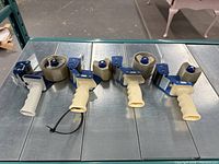 Four handheld packing tape dispensers on metal surface. Each has blue metal frame and beige plastic handle with clear packing tape rolls loaded.