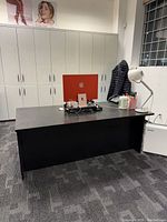 Front angle view of large black wooden desk in office setting showing surface with objects including a laptop, lamp, and other small items, with white cabinets behind.