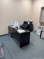 Photo showing black wooden L-shaped desk positioned in office corner, with computer monitor and office chair.