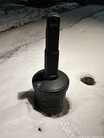 Outdoor cigarette bin standing on snowy ground, viewed from the top side showing the tall cylindrical design and locking mechanism.