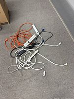 Photo showing multiple extension cords and power strips in a pile on the floor, various colors and sizes visible.