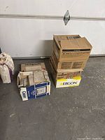 Stack of 4 cardboard boxes with tiles inside, placed on concrete floor near a white wall. Boxes labeled Casagrande and Ergon, one box is open showing beige ceramic tiles inside.