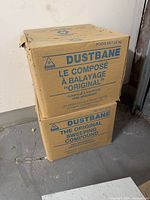 Photo showing two cardboard boxes stacked, labeled Dustbane Original Sweeping Compound with French and English text, placed on floor against wall.