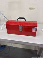 Front view of red rectangular metal toolbox with black handle and silver latch sitting on a white surface.