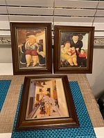 Three framed Botero-style prints, two upright and one laying flat, showing rotund figures in various scenes. Frames are dark brown wood with gold trim.