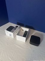 Photo of Apple TV device, remote placed in the packaging tray and original product box on a white surface with a blue background.