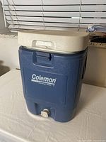 Blue rectangular Coleman cooler with white lid on table, front view showing brand and spigot.