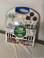 Front view of Dremel 160-piece accessory kit in clear plastic case with blue handle. Label shows $130 value and kit details.