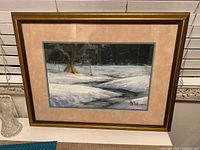 Full view of the framed painting showing a snowy winter landscape with trees, a small creek, and tipis with smoke.
