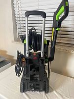 Rear view of electric power washer showing hose reel with black hose coiled, electrical cord coiled, telescoping handle, black and green pistol grip spray gun attached to side.