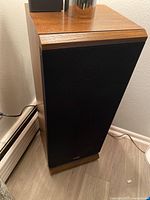 Full view of one speaker showing wooden cabinet and black grille against a wall.