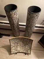 Two tall cylindrical vases with speckled paint and matching curved plate with gold rim on floor and against wall