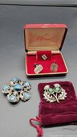 Photo of two brooches on table with a red velvet pouch, one Scottish thistle brooch with green stones, other silver-tone with blue stones and floral pattern.