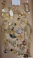 Overview of gold coloured jewelry lot with various necklaces, earrings, a cuff bracelet and pendants laid out on cardboard.