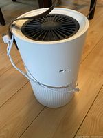 White portable cylindrical air filter with side power button, top vent fan, brown carrying strap, and white USB-C to USB-B cable attached, shown from side angle on wooden floor.