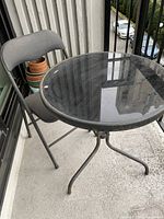 Photo of round glass-top bistro table with folding chair on balcony, showing the table and chair side by side.