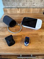 Digital blood pressure monitor with attached arm cuff, blue and white finger pulse oximeter and black carrying pouch on wooden surface.