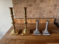 Photo showing two sets of candlesticks on wooden surface against tile wall: two tall polished brass candlesticks with hexagonal bases and two shorter pewter candlesticks with square bases.