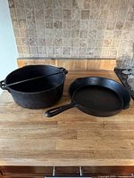 Two black cast iron cooking pieces on countertop: pot with lid, two loop handles, and a skillet with long handle.