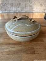 Ceramic serving bowl with lid on wooden surface, beige with speckles and wavy white decoration on lid with ceramic handle