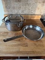 Three stainless steel pots with glass lids, one double boiler steamer insert, and one stainless steel sautee pan shown on wooden kitchen counter.