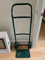 Green metal dolly standing upright against a white wall, showing two black wheels, flat loading plate with scuff marks, and a handle with two crossbars.