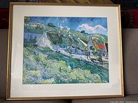 Framed print showing a Vincent van Gogh style landscape featuring cottages with thatched roofs, grassy fields, and blue sky.