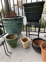 First photo showing large green ceramic pot, black square pot on metal stand, smaller pots of various shapes and colors arranged on balcony floor.