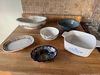 First photo of assortment of 7 serving dishes on wooden countertop including white and patterned ceramic and Pyrex dish with blue cornflower design.