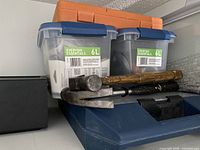 Two plastic storage containers labeled 6L with blue locking lids stacked on top of each other along with three hand tools (a small axe, a hammer, and a block hammer) on a shelf.