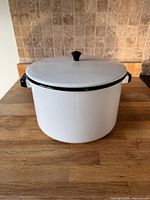 Photo of the vintage white enameled metal pot with lid, placed on wood countertop showing the pot from the side with visible black handles and black knob on the lid.