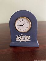 Front view of blue Wedgwood jasperware ceramic clock showing white classical relief decoration and round clock face with Roman numerals